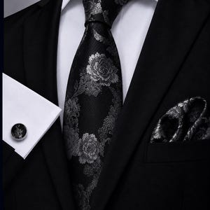 Peut inclure: Une cravate noire ornée d'un motif floral argenté, accompagnée d'une pochette assortie et d'un bouton de manchette rond à bordure argentée. L'ensemble est porté avec une chemise blanche impeccable et une veste de costume noire formelle.