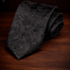 Könnte beinhalten: Eine gerollte schwarze Krawatte mit einem komplizierten Blumen- und Paisley-Muster. Die Krawatte besteht aus einem strukturierten Stoff und wird auf einer dunklen Holzoberfläche präsentiert. Das Design der Krawatte ist in der Nahaufnahme sichtbar.