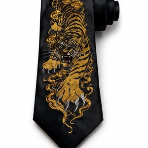 Könnte beinhalten: Eine schwarze Krawatte mit einer detaillierten Stickerei eines Tigers in Gold und Schwarz. Der Tiger ist in einer Sprungposition mit sichtbaren Krallen dargestellt und von wirbelnden, goldfarbenen wolkenartigen Mustern auf dem Stoff umgeben.