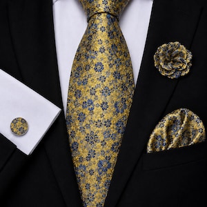 Può includere: Una cravatta dorata con motivo floreale blu, una pochette abbinata, un gemello rotondo e un fiore da risvolto dorato e blu. Questi articoli sono indossati con una camicia bianca e una giacca da abito nera, creando un set coordinato.