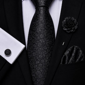 Peut inclure: Une cravate noire avec un motif floral ton sur ton, une épingle de revers fleurie noire assortie, une pochette de costume fleurie noire et un bouton de manchette noir avec une incrustation florale, portés sur une veste de costume noire et une chemise blanche.