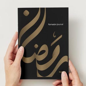 May include: A black journal with gold Arabic calligraphy and the words "Ramadan Journal" printed in white. The journal is held by two hands. The cover features a large, stylized design.