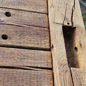 Könnte beinhalten: Nahaufnahme einer verwitterten Holzkonstruktion, möglicherweise ein Tisch oder eine Bank. Das Holz hat ein natürliches, gealtertes Aussehen mit sichtbarer Maserung, Ästen und Löchern. Die Struktur besteht aus dicken, rechteckigen Brettern und Balken.