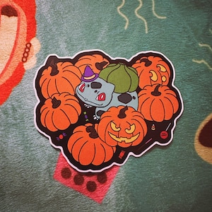 May include: A sticker featuring a green and blue Pokemon character wearing a witch's hat, surrounded by orange pumpkins. The pumpkins are decorated with jack-o'-lantern faces and candy corn.