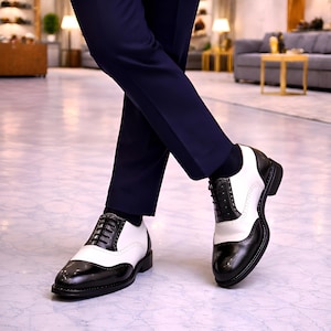 Può includere: Scarpe eleganti in pelle bicolore, nere e bianche. Le scarpe presentano un classico design Oxford con chiusura stringata e suola robusta. Le scarpe sono indossate con pantaloni blu navy.