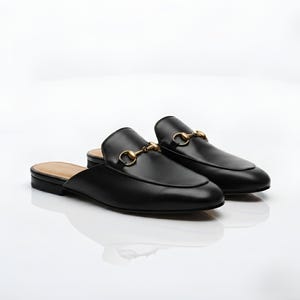 May include: A pair of black leather backless loafers featuring a rounded toe and a gold-toned metal horsebit detail across the vamp. These slip-on shoes have a low heel and a tan-colored interior lining, presented against a plain white background.