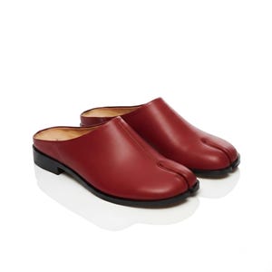 May include: A pair of dark red leather mules featuring a distinctive split-toe design. These slip-on shoes have a smooth finish, a low black heel, and a tan interior lining, showcasing a clean silhouette with a prominent seam down the center.