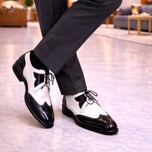 Può includere: Scarpe Oxford bicolore nere e bianche. Le scarpe hanno una finitura lucida sulle sezioni nere, con un motivo decorativo sulla punta e un design unico sul lato. Lacci neri.