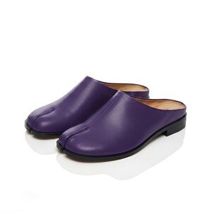 Könnte beinhalten: Ein Paar tiefviolette Ledermules mit einem markanten Zehensteg-Design. Diese Slipper verfügen über eine glatte, matte Oberfläche, einen niedrigen schwarzen Absatz und ein beiges Innenfutter, was eine klare, schlichte Silhouette ergibt.
