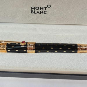 May include: A black and gold Montblanc pen with a red jewel on the clip, displayed in a white box. The pen has gold detailing and a pattern of small gold dots. The Montblanc logo is visible on the box.