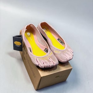 May include: A pair of light pink five-toe shoes with a mesh upper and bright yellow insoles. The shoes feature individual toe compartments, a Vibram logo on the side, and are displayed on a brown cardboard box with a black product tag attached.