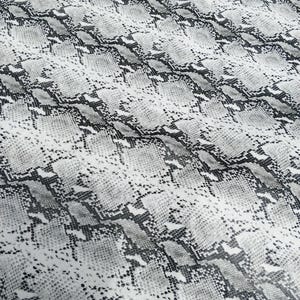 Könnte beinhalten: Ein Textil mit Schlangenmuster. Der Stoff weist ein sich wiederholendes Design aus grauen, schwarzen und weißen Schuppen auf, wodurch ein realistischer Reptilienhaut-Effekt entsteht. Das Material ist drapiert und zeigt die Textur und Flexibilität des Stoffes.