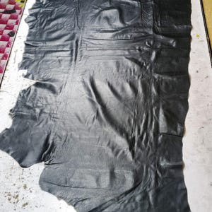 Pode incluir: Uma grande peça de couro preto com formato irregular. O couro tem uma superfície texturizada com vincos e dobras visíveis. O material parece flexível e adequado para a criação de artigos de couro.