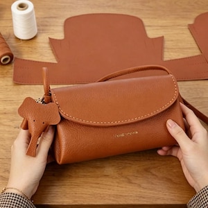 May include: A brown leather crossbody bag with a flap closure and a small elephant charm. The bag has a stitched design and a long strap. The background includes leather pieces and sewing supplies.