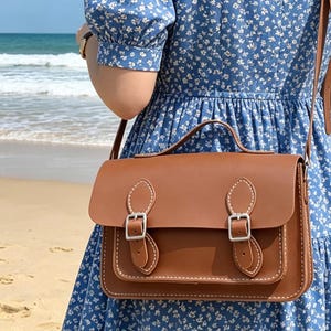 K&ouml;nnte beinhalten: Eine braune Umh&auml;ngetasche mit einem Tragegriff und einem langen Schulterriemen. Die Tasche verf&uuml;gt &uuml;ber eine Frontklappe, die mit zwei silberfarbenen Metallschnallen gesichert ist, sowie wei&szlig;e Kontrastn&auml;hte an den R&auml;ndern der Klappe und Riemen.