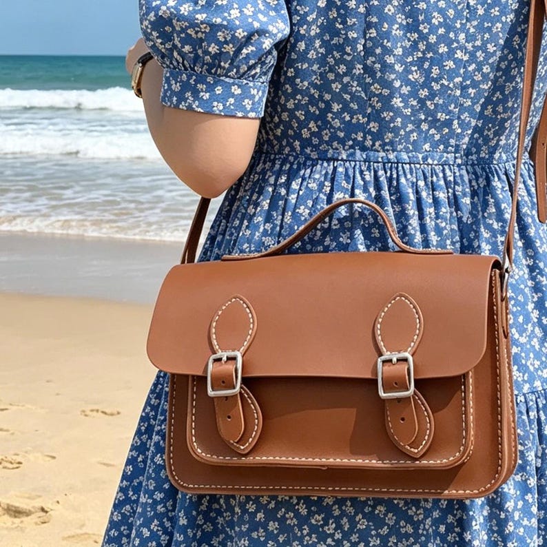 K&ouml;nnte beinhalten: Eine braune Umh&auml;ngetasche mit einem Tragegriff und einem langen Schulterriemen. Die Tasche verf&uuml;gt &uuml;ber eine Frontklappe, die mit zwei silberfarbenen Metallschnallen gesichert ist, sowie wei&szlig;e Kontrastn&auml;hte an den R&auml;ndern der Klappe und Riemen.