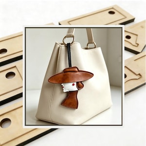May include: A cream-colored handbag featuring a decorative leather charm shaped like a person wearing a wide-brimmed brown hat. The charm has a white face, a red dot accent, and is attached to the bag's gold-toned ring by a thin black strap.