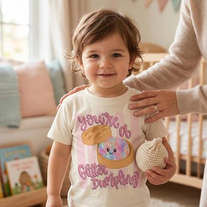 May include: A child wearing a cream-colored t-shirt with the text "You're a Glitter Dumpling" in pink and purple. The shirt features a graphic of a dumpling with glitter and a smiling face. The child is holding a small, beige crocheted ball.