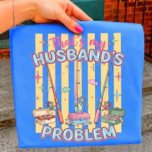 Pode incluir: Tecido quadrado azul com o texto "That's my husband's problem" em rosa e azul. O design inclui varas de pesca, caixas de pesca, peixes e estrelas. O fundo tem listras amarelas e brancas.