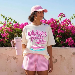 May include: Cream-colored t-shirt with the text "Whatever floats your" and a pink pontoon boat graphic. The person is wearing a pink baseball cap and pink and white checkered shorts. A pink tumbler is held in the hand.