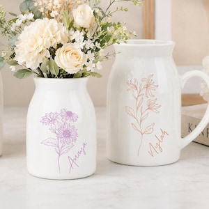 May include: Three white ceramic pitchers with handles, each featuring a delicate line-art floral illustration and a name in cursive script. The left pitcher has a yellow flower, the center has a purple flower, and the right has a pink flower.