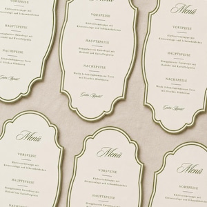 Könnte beinhalten: Eine Sammlung von cremefarbenen Menükarten mit einem verzierten, geschwungenen Rand und einer dünnen olivgrünen Umrandung. Jede Karte zeigt oben das Wort Menü, gefolgt von den Abschnitten Vorspeise, Hauptspeise, Nachspeise und dem Text Guten Appetit.