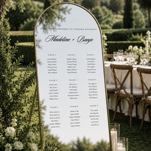 Peut inclure: Un panneau de plan de table de mariage en forme d'arche avec un cadre en métal doré, affichant le texte "HERZLICH WILLKOMMEN ZU UNSERER HOCHZEIT", les noms "Madeline + Banjo" et une liste d'invités organisée par neuf tables sur fond blanc.