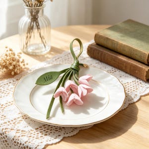 May include: A decorative keychain featuring a cluster of soft pink fabric flower blossoms attached to a green leather loop. One green leather leaf is attached to the stem and features the name Mia embossed in a dark, clear, sans-serif font.