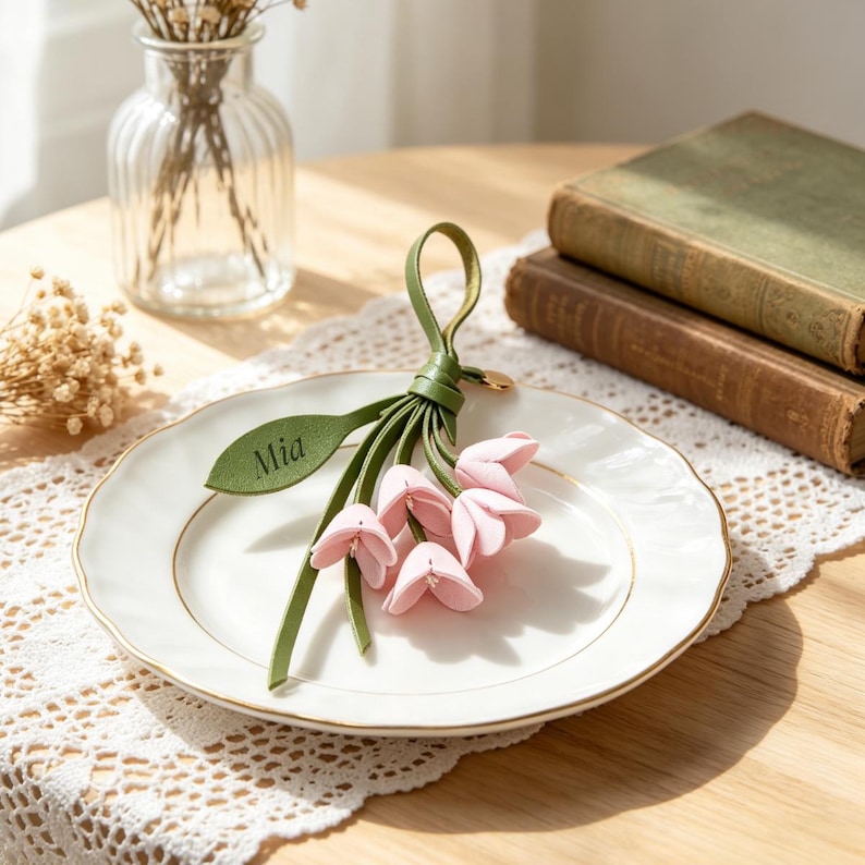 May include: A decorative keychain featuring a cluster of soft pink fabric flower blossoms attached to a green leather loop. One green leather leaf is attached to the stem and features the name Mia embossed in a dark, clear, sans-serif font.
