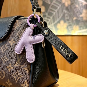 Può includere: Primo piano di una borsa marrone e nera con un portachiavi a forma di lettera "K" in pelle lavanda e un cinturino in pelle nera con il nome "LUNA" in lettere dorate. Il portachiavi è attaccato al manico della borsa con un anello dorato.