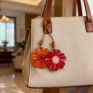 Op de afbeelding: Een beige handtas met bruine leren handvatten en accenten. Twee leren bloembedels in oranje en rood hangen aan het handvat. De bloemen hebben gedetailleerde bloemblaadjes en zijn bevestigd met geknoopte leren koorden.