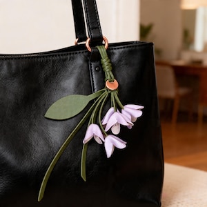 May include: A black leather tote bag featuring a decorative charm attached to the handle. The charm consists of three light purple bell-shaped flowers with green stems and a single green leaf, all secured by a copper-colored metal ring.