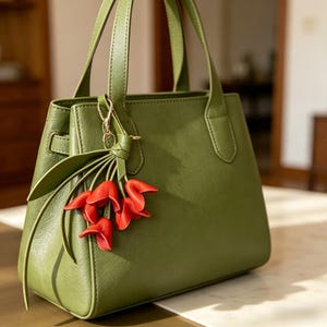 May include: A structured olive green handbag with two top handles and a gold-toned metal clasp. A decorative charm featuring several small, bright red flower-shaped pendants hangs from one of the handles, adding a distinct accent to the bag.