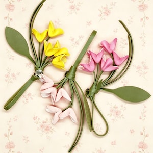 May include: Two decorative floral ornaments crafted from green leather-like material. One features bright yellow and pale pink petals, while the other displays vibrant pink petals. Both include long green stems, leaves, and a circular metal fastener.