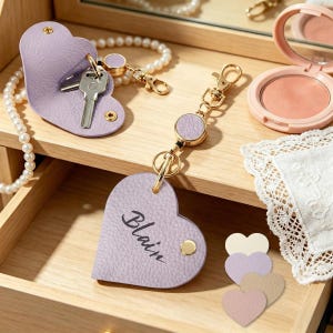 Puede incluir: Dos llaveros de cuero en forma de corazón de color lavanda con herrajes dorados y carretes retráctiles. Un llavero está abierto mostrando una llave de metal, mientras que el otro está cerrado y tiene el nombre Blain impreso en negro.