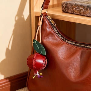 Può includere: Una borsa a spalla in pelle marrone con cerniera dorata e un ciondolo a forma di melograno. Il ciondolo è realizzato in pelle rossa con cuciture a vista, una foglia in pelle verde e piccoli accenti gialli e viola alla base del frutto.