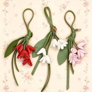 May include: Three decorative hanging ornaments shaped like flower bouquets, each featuring a green leather loop and stem with petals in red, white, and light pink. The ornaments are arranged vertically, showcasing detailed floral shapes and long green leaves.