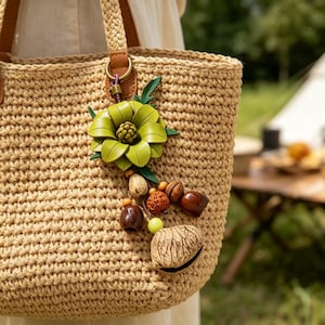 May include: Woven straw tote bag with brown leather handles and a decorative green flower and beaded charm. The bag features a braided handle and a light brown color. The charm includes wooden beads and a shell.