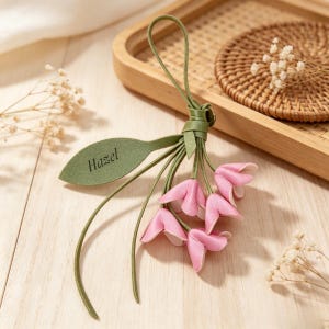 May include: A decorative keychain featuring a cluster of small, light pink bell-shaped flowers attached to a green stem and a single green leaf. The leaf is engraved with the name Hazel in a dark script font. The item includes a green loop for hanging.