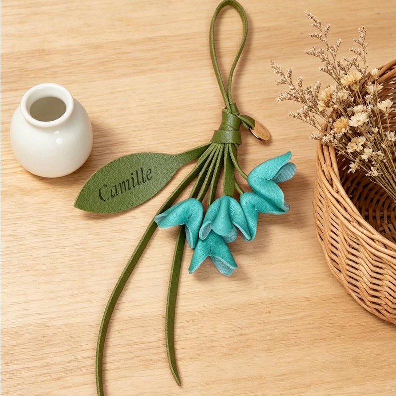 May include: A decorative hanging ornament featuring a green leather loop and a matching green leaf-shaped tag with the name Camille embossed in black. Attached are several teal-colored, bell-shaped leather flower petals hanging from a central knot.