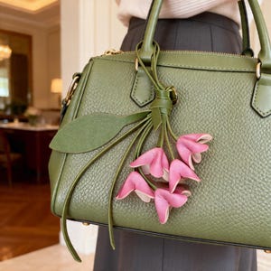May include: An olive green textured leather handbag featuring a top zipper closure, two sturdy handles, and a decorative charm consisting of a green leather leaf and a cluster of five small, folded pink leather flower petals hanging from a knot.