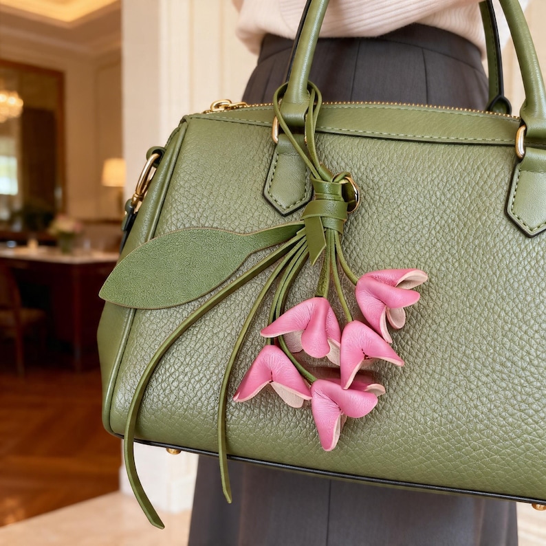 May include: An olive green textured leather handbag featuring a top zipper closure, two sturdy handles, and a decorative charm consisting of a green leather leaf and a cluster of five small, folded pink leather flower petals hanging from a knot.