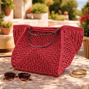 Può includere: Una borsa tote rossa all'uncinetto con manici in metallo nero è appoggiata su un tavolo di pietra. Occhiali da sole tartarugati e braccialetti dorati sono accanto alla borsa. Un cappello di paglia è parzialmente visibile sullo sfondo.