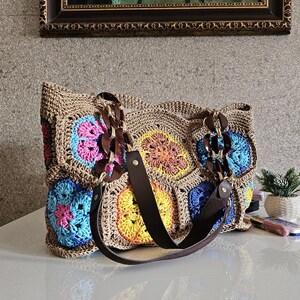 Pode incluir: Uma mala tote de crochê feita à mão com base marrom e padrões florais coloridos em rosa, azul, amarelo e laranja. A mala apresenta alças e detalhes em couro marrom, e um detalhe de corrente. A mala está sobre uma superfície branca.