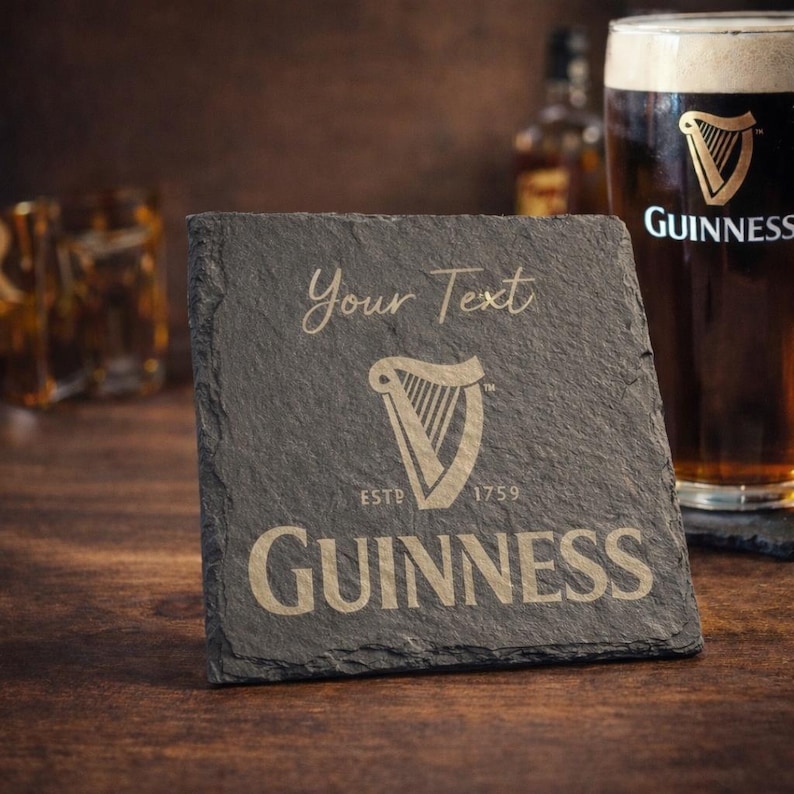 May include: A dark gray slate coaster with the Guinness logo, a harp, and the text "Your Text". The coaster also features the text "ESTD 1759" and the Guinness brand name. A pint of Guinness is in the background.