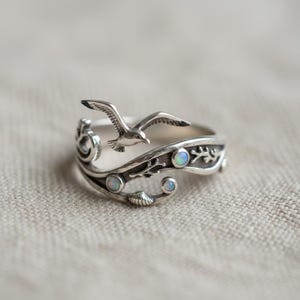 May include: A silver ring with a seagull design, incorporating wave and seashell motifs. The ring includes three small, round, opalescent stones. The design is detailed, with a nautical theme.
