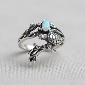 May include: A silver ring featuring a sea turtle design. The ring has an oval, iridescent opal gemstone and small accent stones. The turtle is detailed with a textured shell and flippers, and the ring band has a branch-like design.