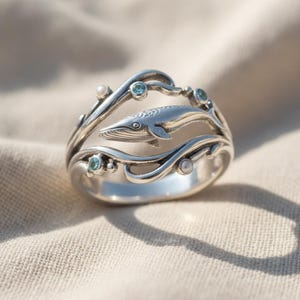 May include: A silver ring featuring a sculpted whale swimming between two curved bands that mimic ocean waves. The design is accented with small, round, light blue gemstones and tiny white pearls embedded along the silver wave structures.