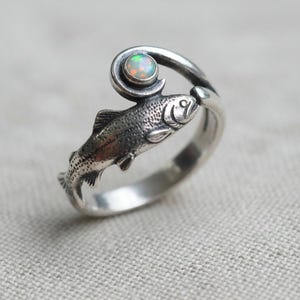 May include: A silver ring with a fish design. The ring features a swirling detail with a small, round opal stone. The fish is textured with scales and fins. The ring wraps around the finger, a unique piece of jewellery.