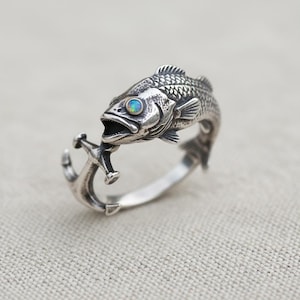 May include: A silver ring featuring a detailed fish design with an open mouth, holding an anchor. The fish has an opal-like stone for an eye. The ring is made of a shiny, metallic material.